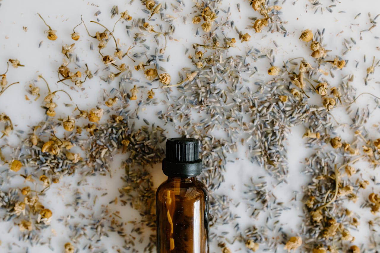 A close-up of an essential oil bottle surrounded by dried lavender and chamomile on a white surface.